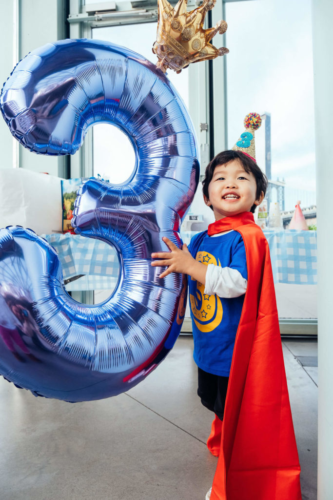 Boy dressed as a superhero holding a large number 3 balloon for a birthday celebration.
