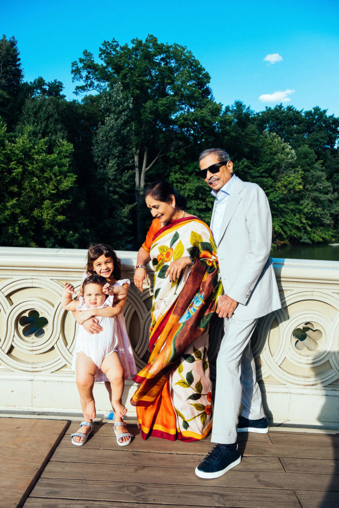 A smiling family of four stands on a bridge with ornate railings in a park, perfectly captured by a Brooklyn family photographer. An elderly couple, with the woman in a colorful saree and the man in a light suit, accompanies two young children. Trees and a blue sky serve as their picturesque backdrop.