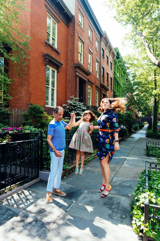 A lively scene on a sunny day: three people joyfully jump while holding hands on a sidewalk lined with brick buildings and greenery, capturing the essence of family photography in NYC. The group consists of two adults—a man and a woman—and a child in a dress, all smiling and carefree.