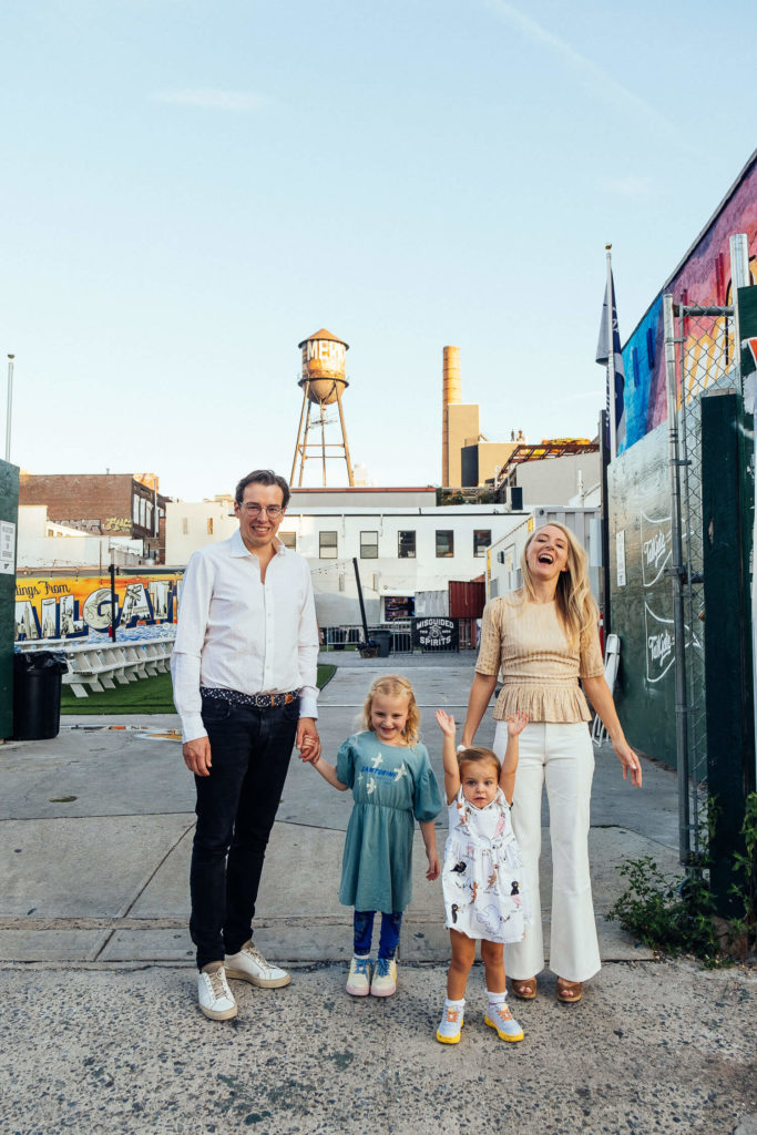 A family of four stands smiling outdoors on a Brooklyn street, captured by a talented Brooklyn family photographer. The parents are on either side, with a young girl and toddler between them. A water tower and industrial buildings set the scene under the clear blue sky, perfect for NYC family photography.