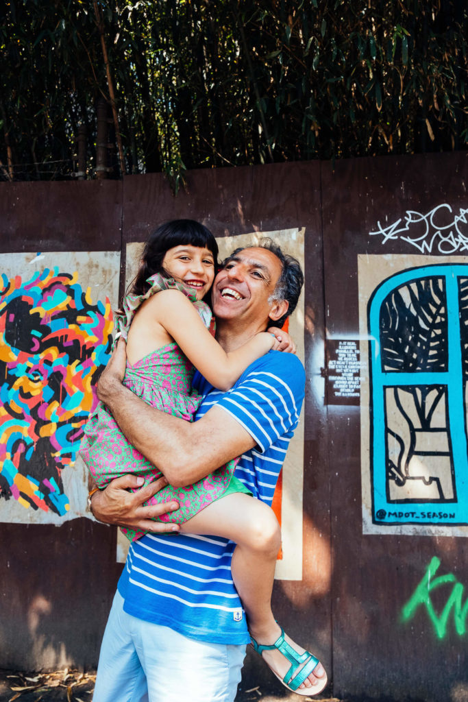 A man in a blue striped shirt joyfully holds a young girl in a colorful dress as they smile in front of vibrant street art. The backdrop, filled with abstract and geometric patterns, captures the essence of family photography NYC, reminiscent of moments captured by a Brooklyn family photographer.