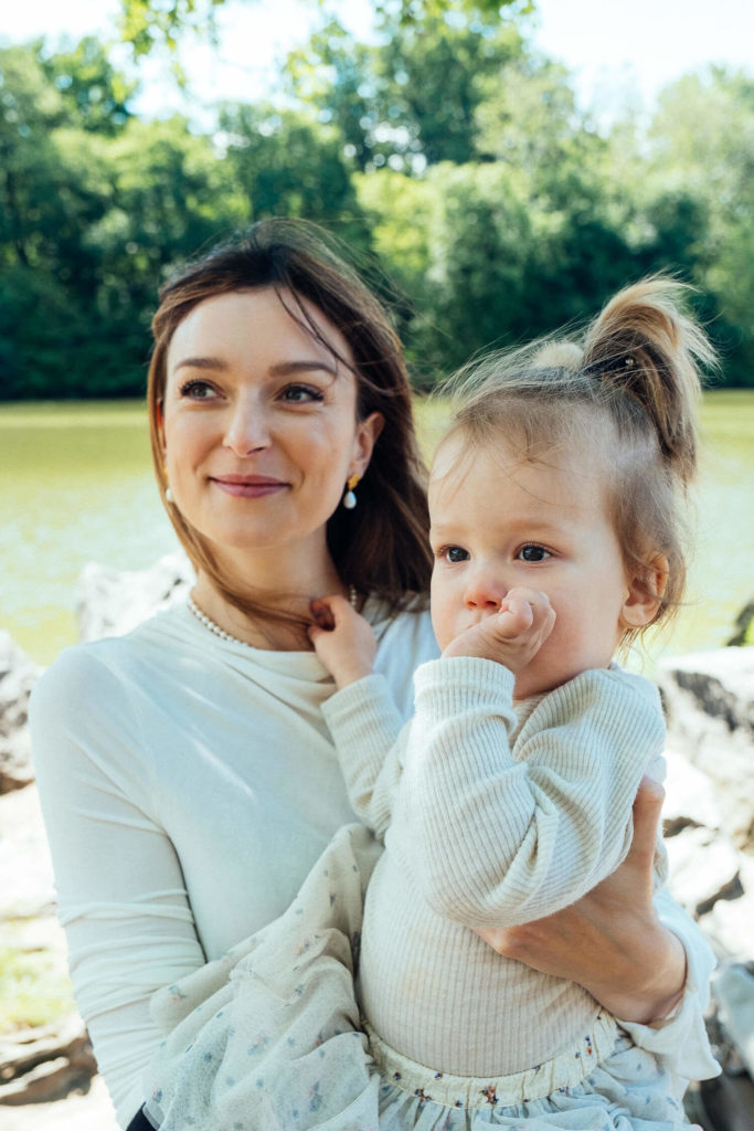 A woman cradles a child by the lake, both dressed in light attire. The little one curiously has a finger in their mouth. With trees and sparkling water beneath a clear sky, this scene captures the essence of family photography NYC style.