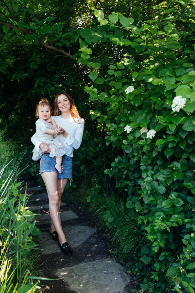 A woman holding a young child stands on a stone path surrounded by lush greenery, captured beautifully by a Brooklyn family photographer. They are both smiling, dressed in casual summer clothes, with white flowers blooming on bushes and sunlight filtering through the leaves.