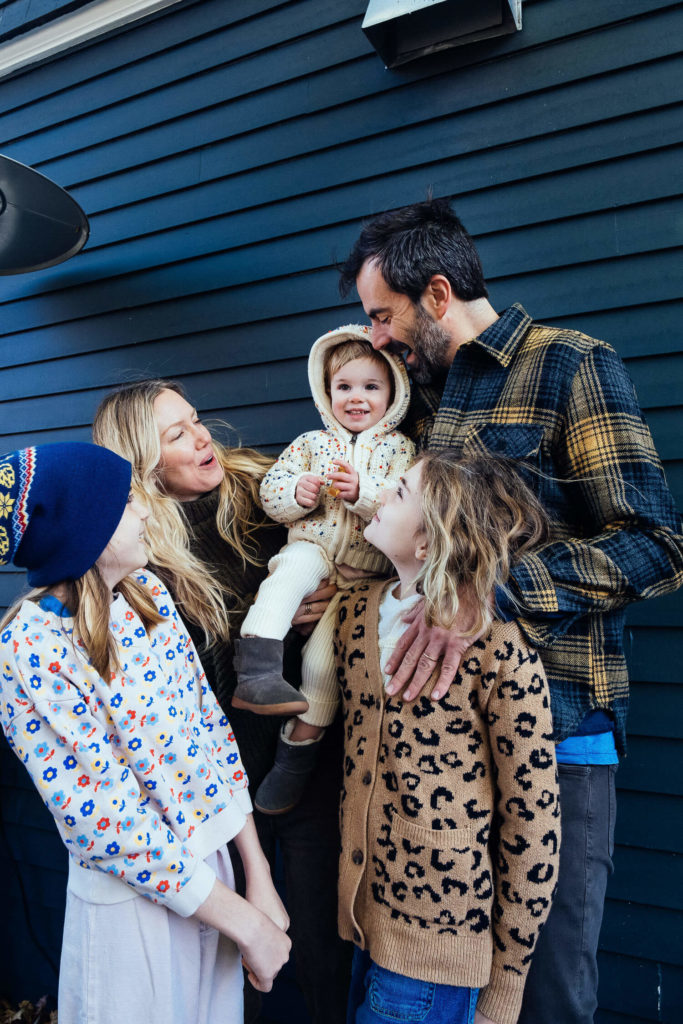 A family of five smiles and gathers closely in front of a dark blue wall, perfectly captured by a talented Brooklyn family photographer. The father holds a toddler in a hooded outfit, while the mother and two children, wearing patterned sweaters, look on affectionately. The atmosphere is joyful.