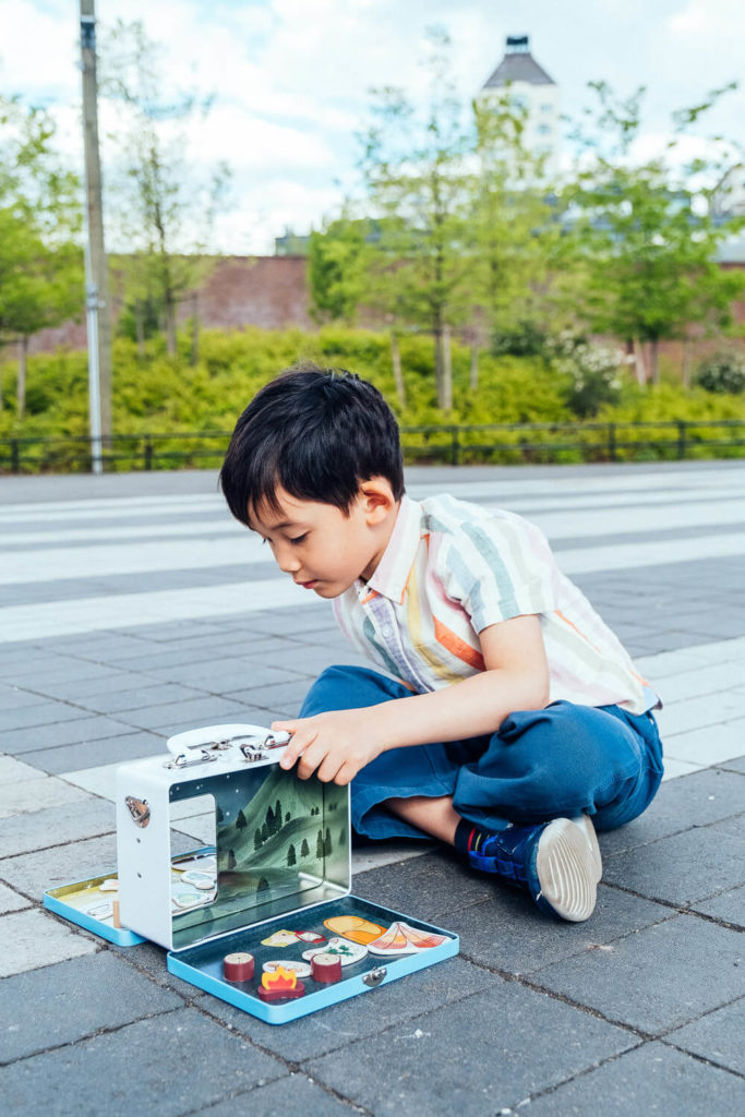 A young boy sits cross-legged on a paved surface, absorbed in play with a miniature kitchen set—a scene perfect for brand photography. Toy food and kitchen items scatter around him, while trees and a building stand peacefully in the background beneath a partly cloudy sky.