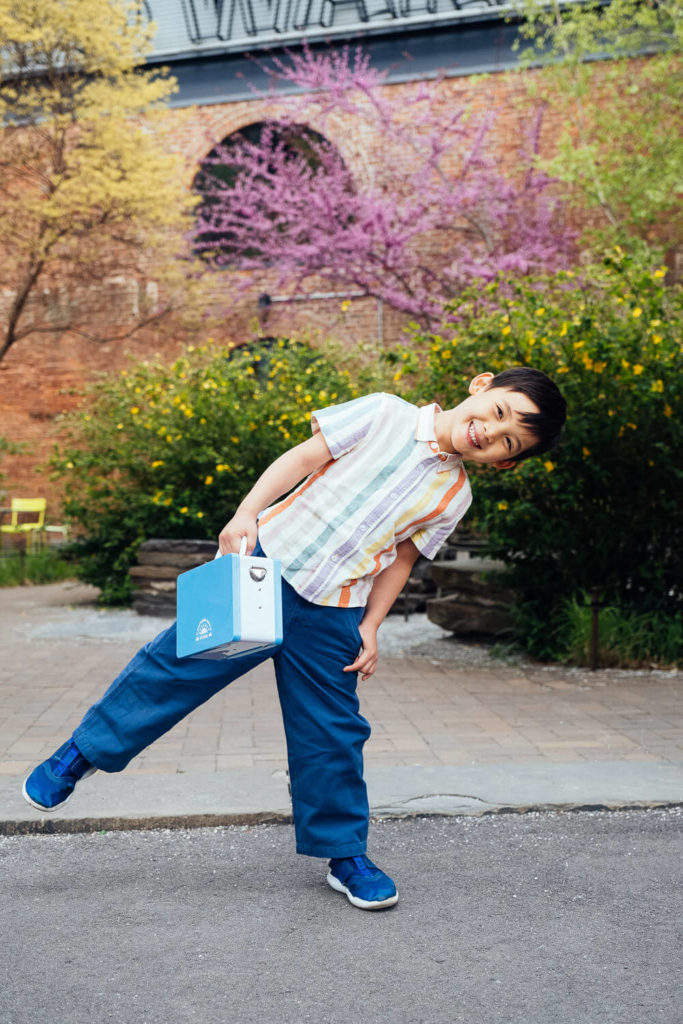 A young boy poses playfully outdoors, embodying the charm of brand photography as he leans to one side with a blue paper bag. Dressed in a striped shirt, blue pants, and shoes, he stands before blossoming trees and a brick building. The setting is bright and cheerful.