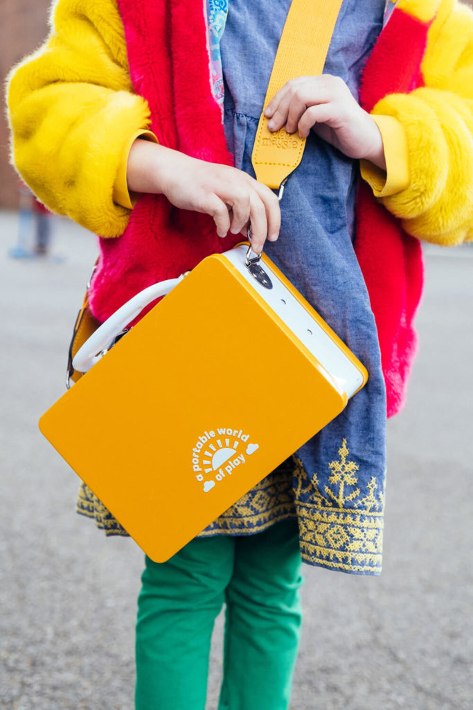 A child in a bright yellow jacket, red vest, and green pants is holding a yellow book with the text 