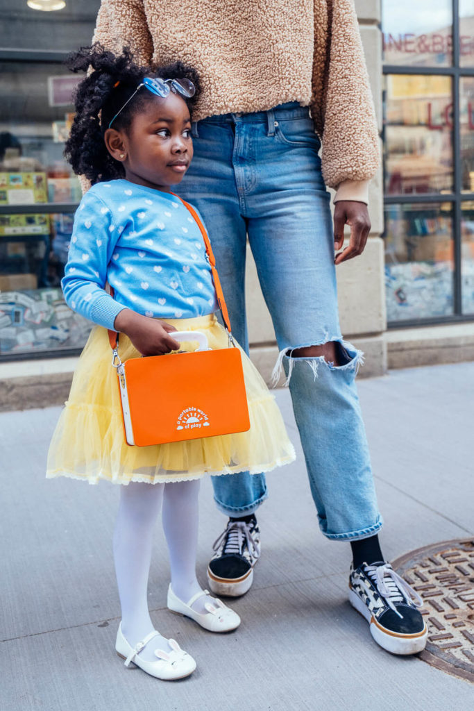 A young girl in a blue sweater and yellow tutu stands with a woman in a brown sweater and ripped jeans, embodying vibrant brand photography. The girl holds an orange bag, wearing white tights and shoes as they pose on a bustling city sidewalk.