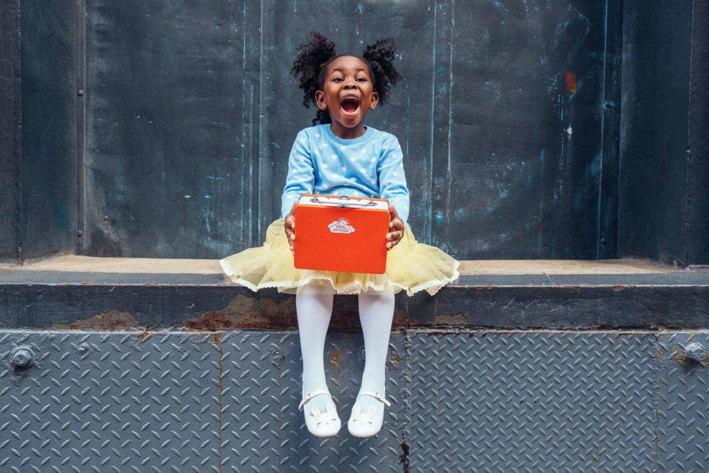 A joyful young girl with curly hair, clad in a blue sweater and yellow skirt, sits on a ledge, embodying the essence of brand photography. She holds a red box, laughter spilling from her open mouth and sparkling eyes against the textured dark wall.