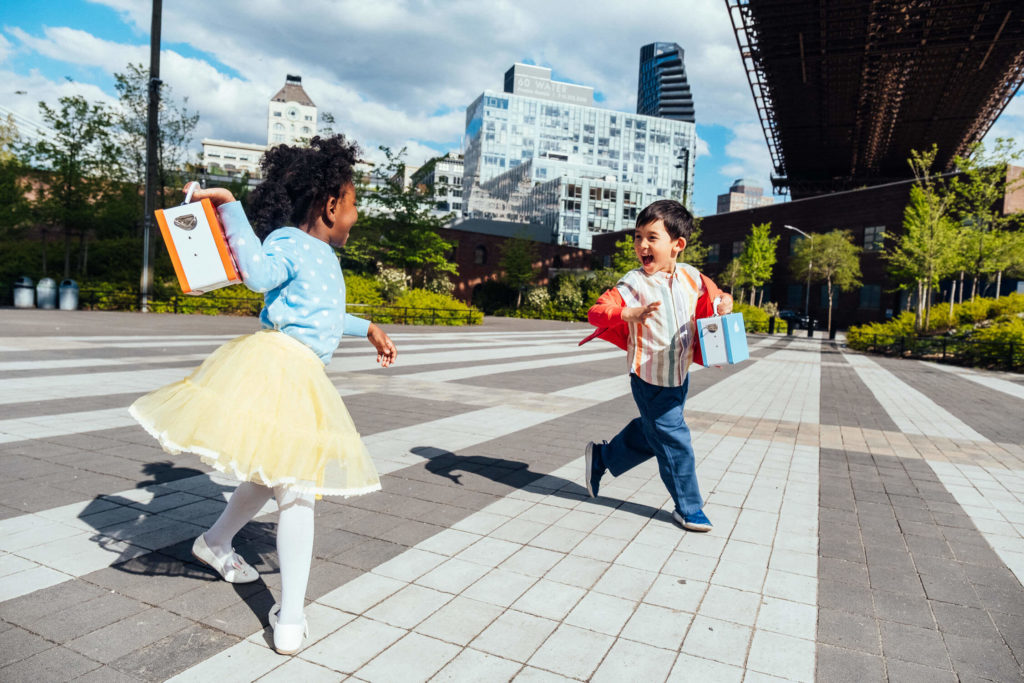 Two children joyfully run across a spacious plaza, each holding a colorful book. The sunny day highlights modern city architecture and greenery in the background, creating a lively and vibrant atmosphere perfect for brand photography.