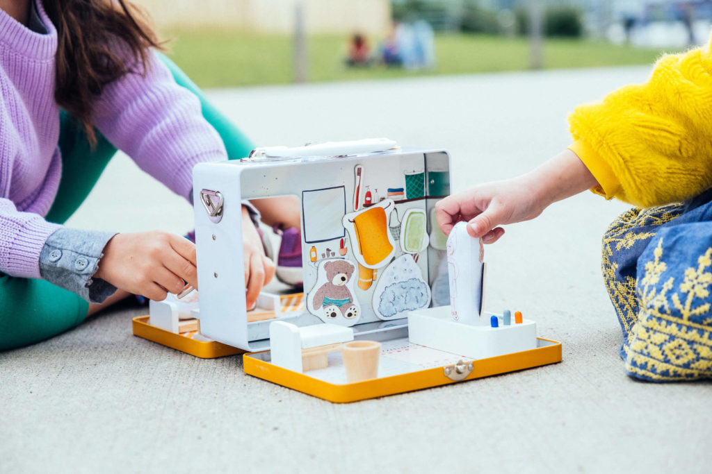 Two children are playfully engaged with a vibrant wooden toy kitchen set on the ground, perfect for brand photography. The kitchen set stands out in white and yellow, accompanied by colorful accessories. The children, dressed in purple and yellow outfits, add a lively touch to this commercial photography scene.