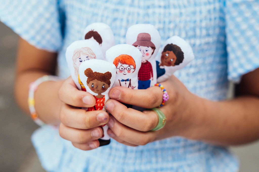 A child in a blue checkered shirt holds several small fabric dolls, each featuring a cartoon character with varying hairstyles and clothing. The child's hands, adorned with colorful rings and bracelets, exude charm in this delightful piece of brand photography.