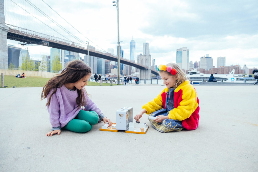 Two children, in a scene perfect for brand photography, play a tabletop game on a concrete surface in the park. One sports a purple sweater and green pants, while the other wears a colorful jacket, with the Brooklyn Bridge and city skyline painting an iconic backdrop.