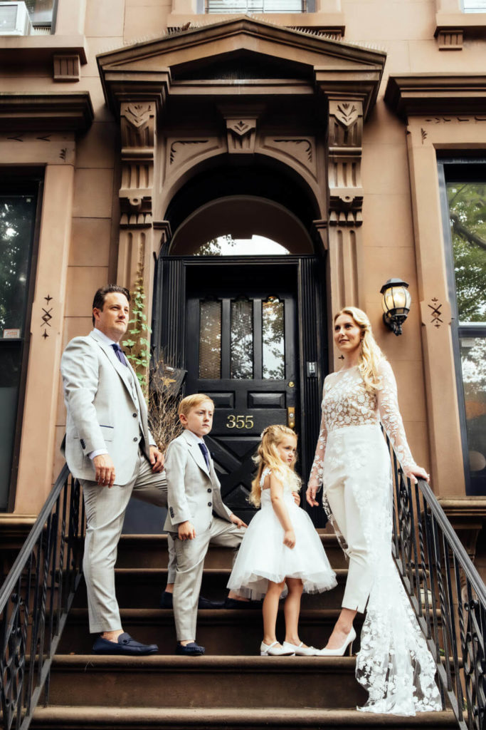 A family of four poses on the steps of a brownstone building, a classic subject for any Brooklyn family photographer. The parents wear formal attire, with the mother in a white lace gown. Their young son and daughter, also elegantly dressed, stand between them by the door numbered 355.