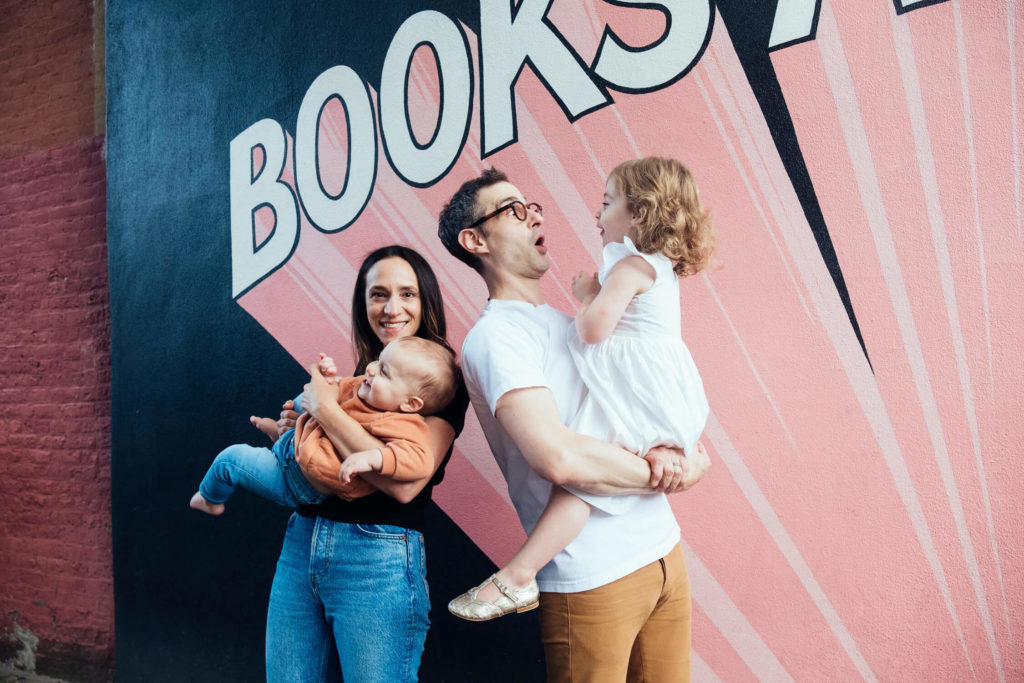 A family of four stands joyfully against a colorful mural that says BOOKS. Captured by a talented Brooklyn family photographer, the mother holds a baby as the father lifts a young girl in a white dress. They all smile warmly in their casual attire, surrounded by vibrant pink and dark hues.