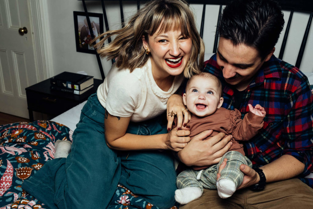 A joyful family moment on a bed features a smiling woman, a man holding a laughing baby, and a quilt with colorful patterns. Captured by a Brooklyn family photographer, the scene conveys warmth and happiness, with books visible on a table in the background.