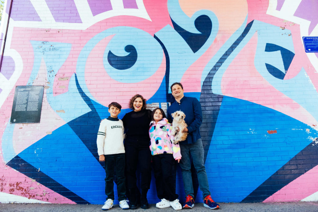 A family of four, with two children and a small dog, stands smiling in front of a colorful mural on a brick wall. Captured by a Brooklyn family photographer, the mural features abstract shapes in shades of pink, blue, and orange—a perfect spot for family photography NYC moments.