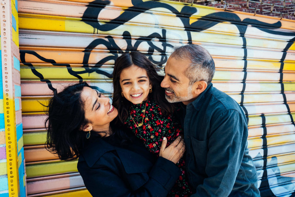 A family of three stands joyfully in front of a colorful graffiti-covered wall, captured by a renowned Brooklyn family photographer. The woman and man smile warmly at their daughter, who is in the center wearing a floral dress and grinning. The atmosphere exudes happiness and relaxation.