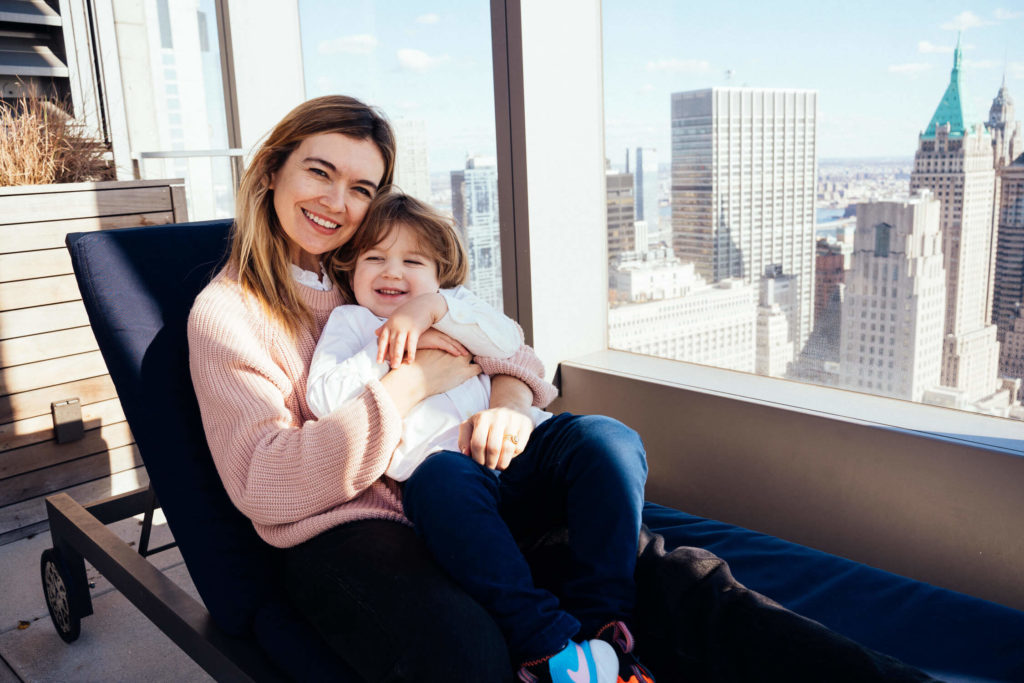 A woman sits on a lounge chair, smiling and holding a young child on her lap. Capturing this perfect family photography moment in NYC, they enjoy their rooftop view of towering city buildings under a clear blue sky. The woman wears a pink sweater, while the child sports a white shirt and blue pants.