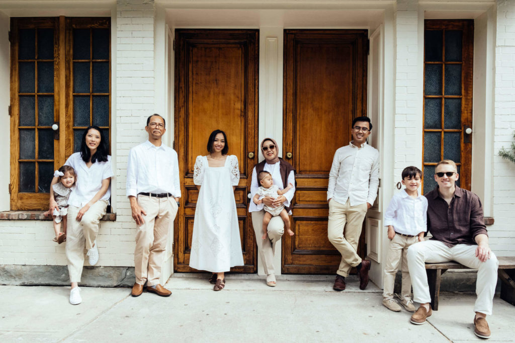 A diverse group of eight, including men, women, and two young children, pose in front of two wooden doors. Some stand while others sit, all in light-colored outfits on a bright day. The scene captures the warm and friendly atmosphere typical of family photography NYC sessions.