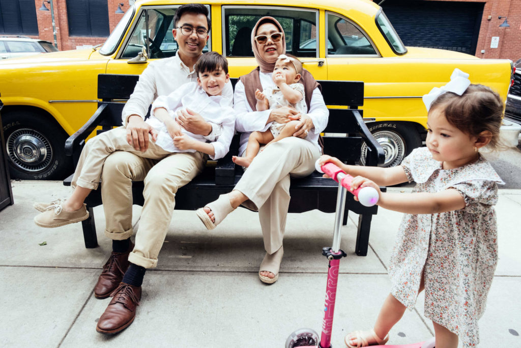 A family sits on a bench with a yellow taxi in the background, embodying quintessential family photography NYC offers. A man and woman hold a baby and a child, while another child stands nearby on a scooter in a floral dress and bow, capturing an urban street moment.