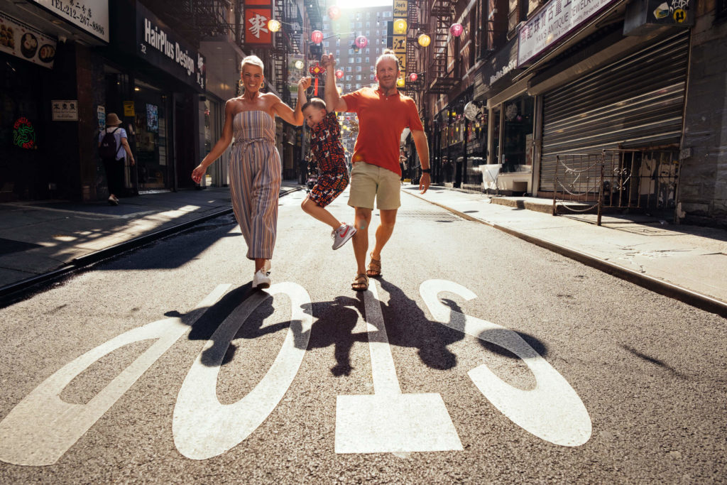 A family of three joyfully walks hand-in-hand down a sunny street, perfectly capturing the essence of family photography in NYC. The child playfully swings between the parents as colorful paper lanterns and shopfronts decorate the background, reminiscent of a Brooklyn family photographer's dream scene.