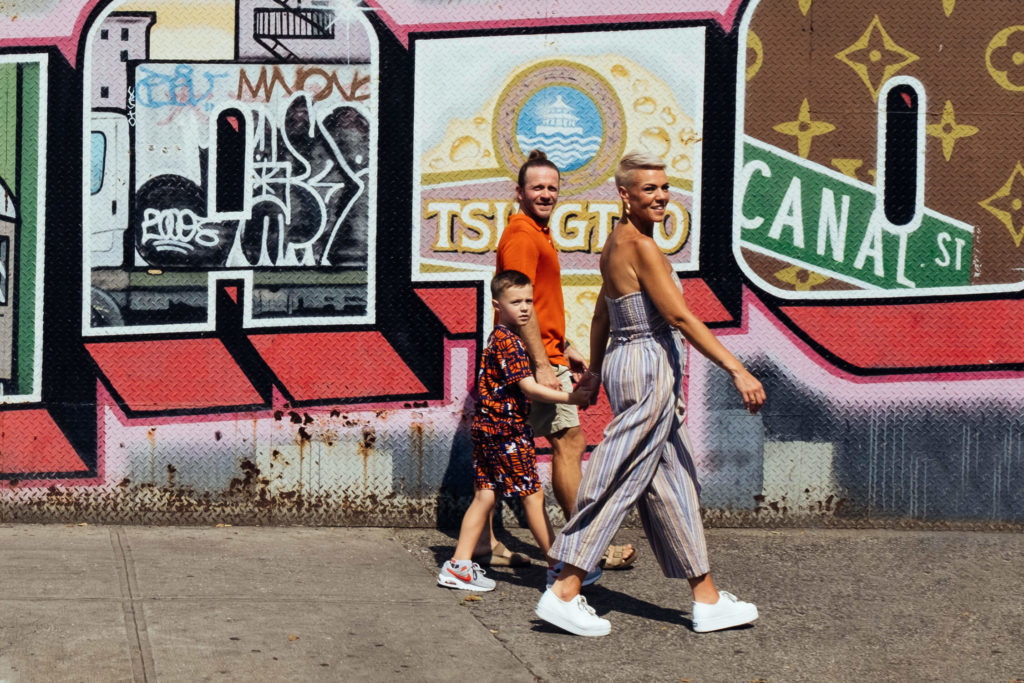 A family of three, including a child, strolls past a colorful street mural in NYC. Captured by a Brooklyn family photographer, the mural bursts with vibrant pink, orange, and green hues while the sun casts long shadows on the sidewalk.