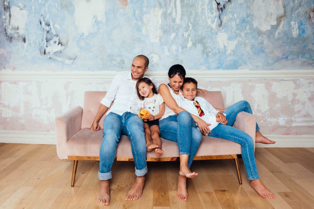 A family of four sits on a light pink couch against an abstract painted wall, capturing a perfect moment of joy with a Brooklyn family photographer. The parents and two children are smiling in their casual clothes, as the mother hugs her daughter, who holds onto her favorite toy.