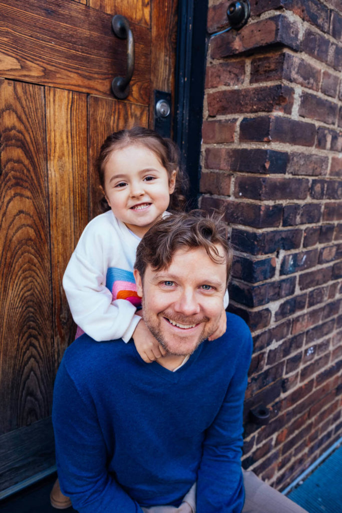 A child and an adult smile while posing outside a brick building. The child, in a colorful sweater, wraps their arms around the adult's shoulders. The adult, in a blue sweater, sits on a step in front of a wooden door—a beautiful family photography moment captured by a Brooklyn family photographer.