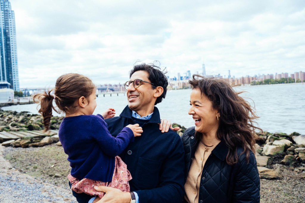 A happy family of three is captured by a waterfront with a city skyline in the background, reminiscent of family photography NYC loves. The father holds their young girl in a purple cardigan, while the mother smiles, all enjoying the cloudy day outdoors.