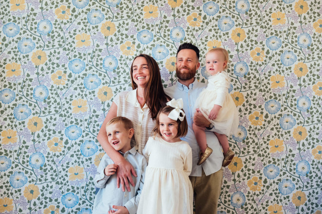 A smiling family of five stands together in front of a colorful floral wallpaper, captured beautifully by a Brooklyn family photographer. The parents hold an infant, while two young children stand in front, all wearing light-colored clothing—a perfect snapshot of family photography in NYC.