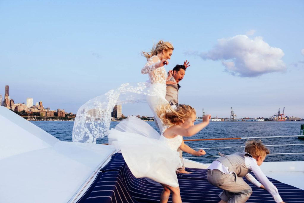 A joyful bride and groom, in wedding attire, leap playfully off a boat's deck. Two children in formal clothes jump beside them. Captured by a Brooklyn family photographer, the backdrop is a city skyline under clear blue skies, reflecting the essence of family photography in NYC.