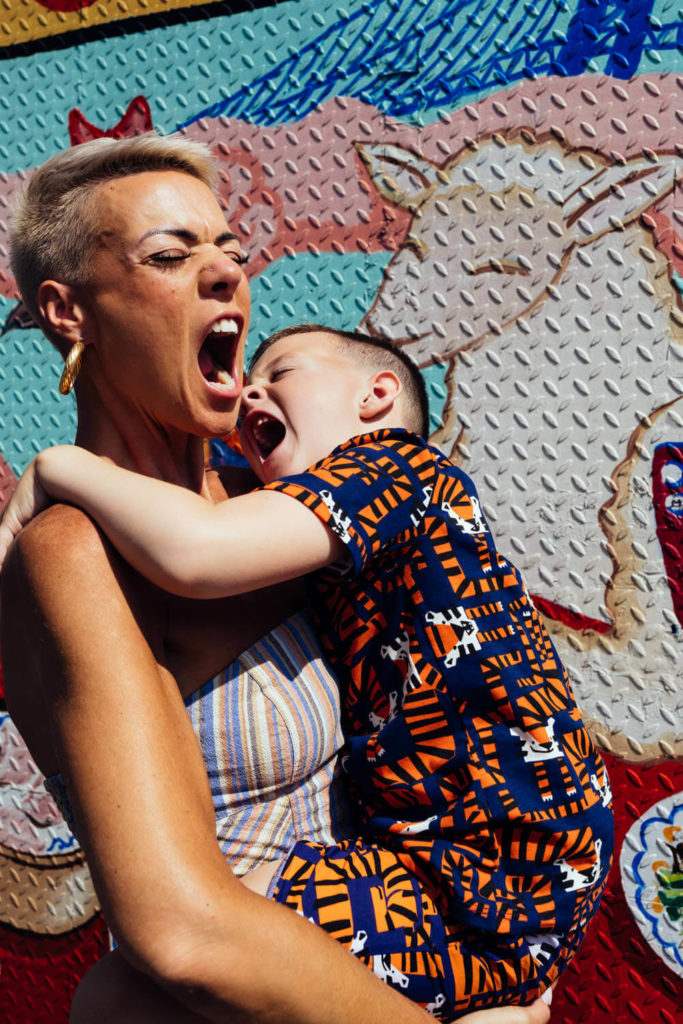 A person with short hair holds a playfully yelling child in front of a textured, colorful mural featuring a llama. The child wears an outfit with orange and blue patterns. Captured by a Brooklyn family photographer, both appear to be enjoying the moment in this vibrant slice of family photography NYC.