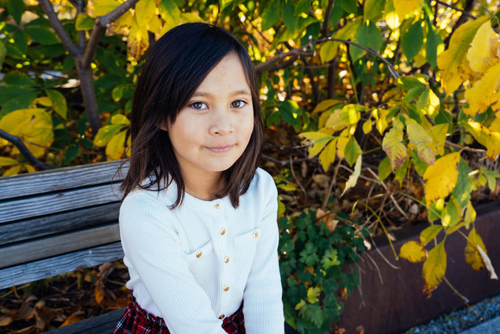 Girl posing in front of a wall decorated with red heart illustrations, smiling upwards.
