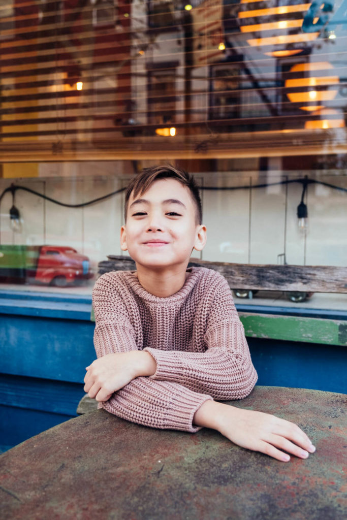 boy smiling while leaning on a table in front of a storefront, relaxed and casual atmosphere