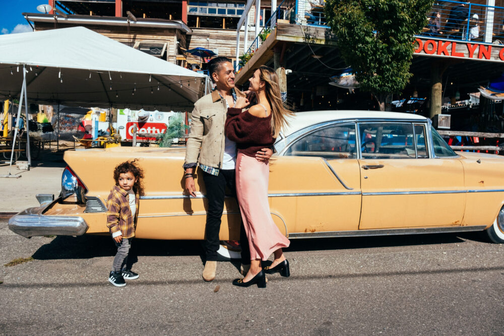 A man, woman, and young child stand smiling together in front of a vintage yellow car on a sunny day, with colorful buildings and tents in the background.