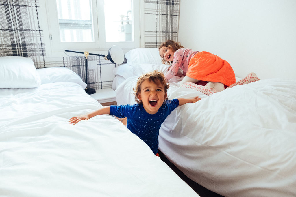 Two young children play and laugh on white beds in a bright, cozy bedroom. One child in blue stands between the beds, arms outstretched and smiling, while the other child in orange plays on the bed in the background.