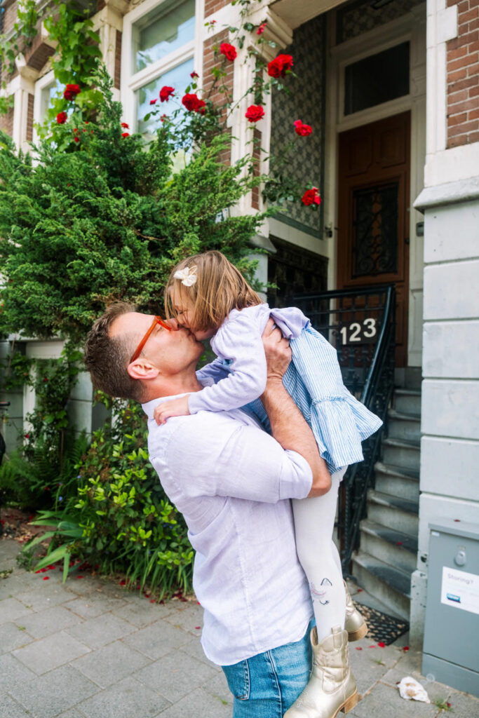 A man in a light shirt lifts a young girl in a blue dress as they share a kiss, captured beautifully by a Brooklyn family photographer. They stand on a sidewalk before a brick building with roses climbing near the entrance and steps leading to the door.
