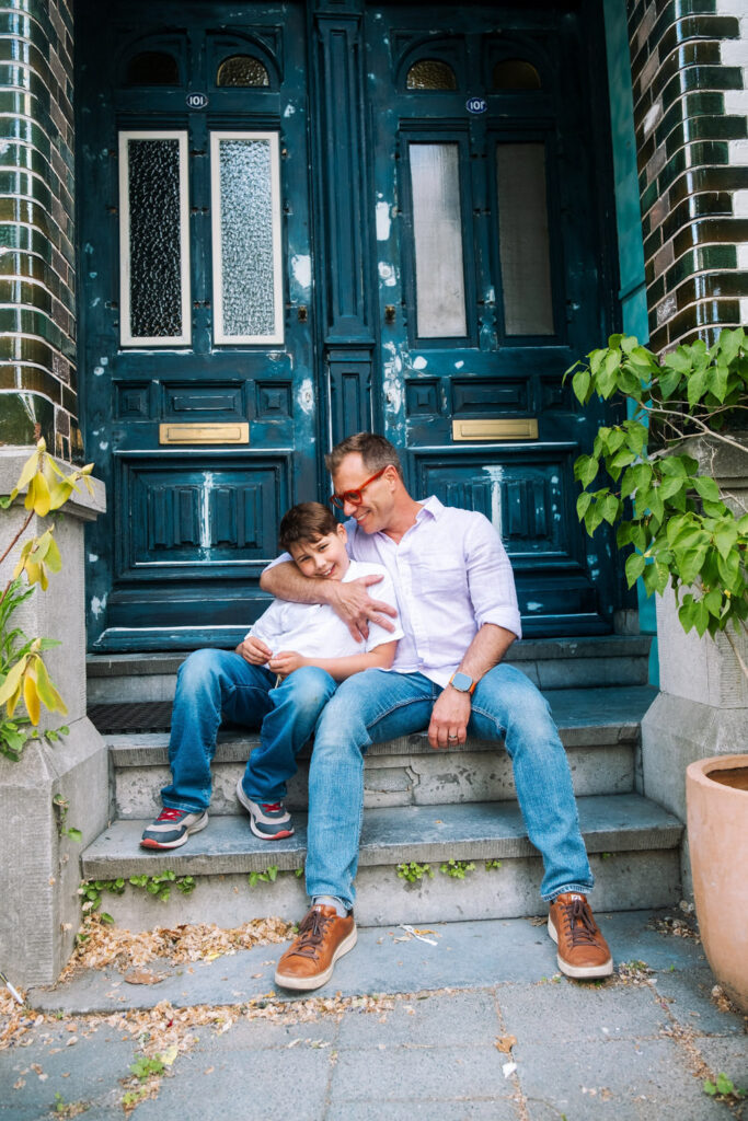 A man and a boy sit on stone steps in front of a blue double door, smiling and hugging. They both wear casual clothes and glasses, enjoying a happy moment together—perfect for family photography NYC style.