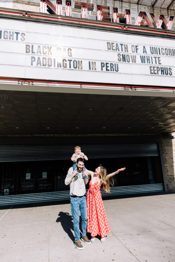 A man holds a child on his shoulders while a woman in a red checked dress gestures excitedly outside a vintage theater with a marquee overhead—an unforgettable moment captured by a Brooklyn family photographer.