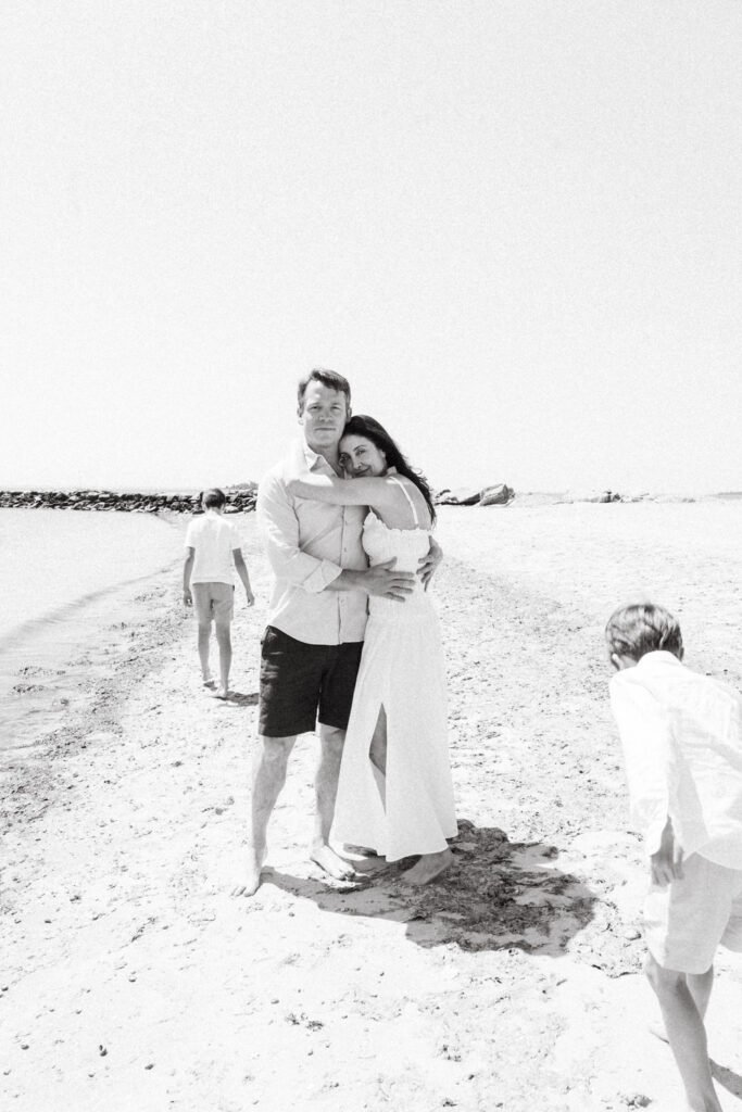 A man and woman embrace on a sandy beach, dressed in light clothing, while two children play nearby. Captured by a Brooklyn family photographer, the bright scene features calm water, rocks, and warm sunlit moments perfect for family photography NYC.