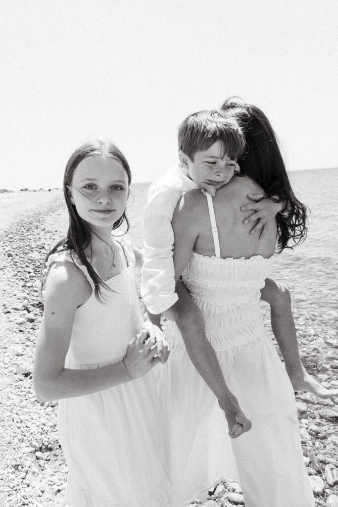 A black and white photo of a young girl in a white dress holding hands with a woman, who carries a boy on her hip. Captured by a Brooklyn family photographer, they stand on a rocky beach with the ocean in the background.