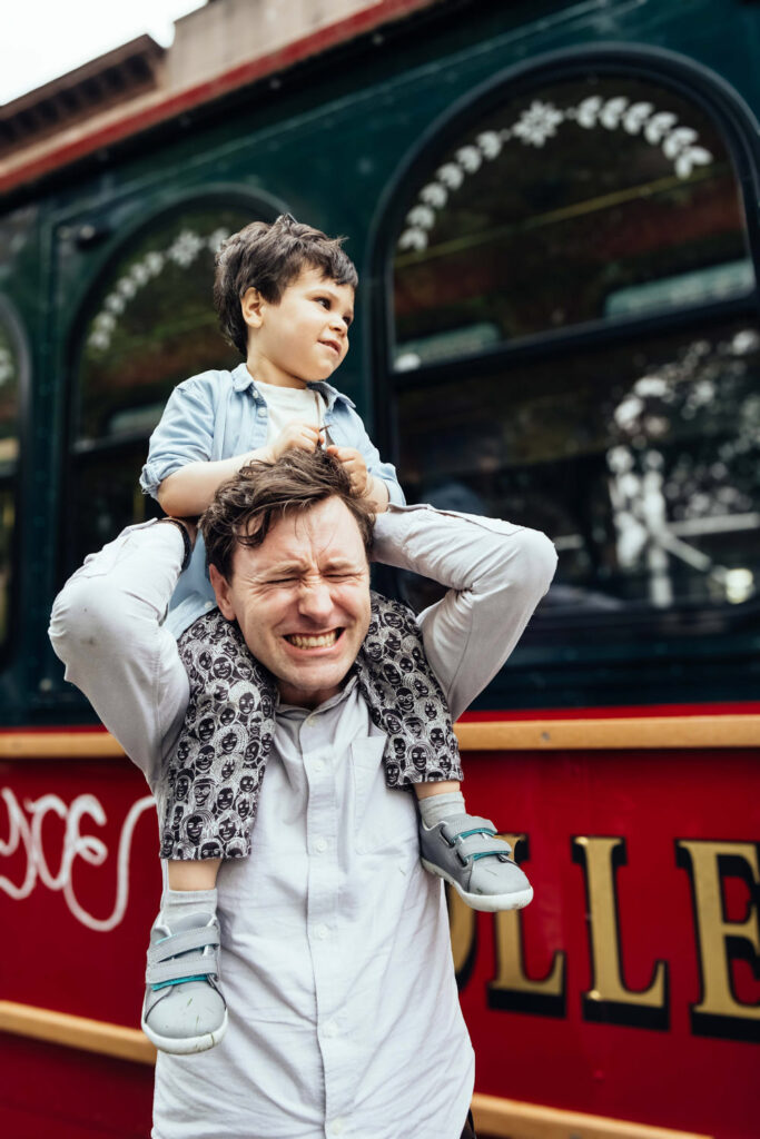 A man smiles with his eyes closed as he carries a young boy on his shoulders in front of a red trolley. Captured by a Brooklyn family photographer, the boy looks off to the side, appearing curious while holding onto the mans head for balance.