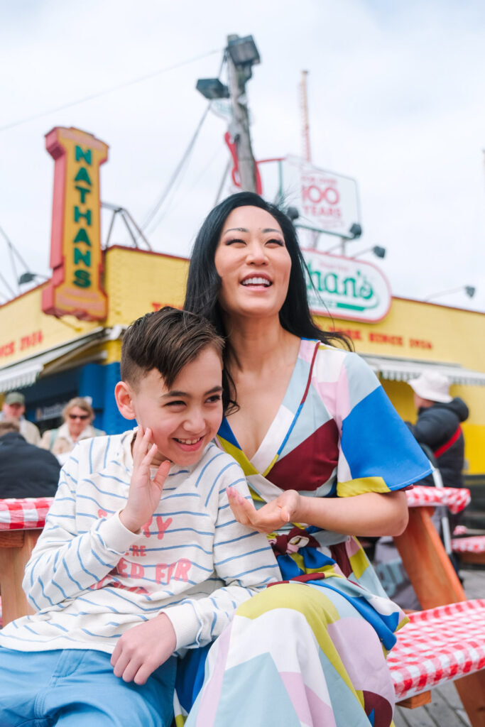 A woman in a colorful dress sits at an outdoor table with a smiling young boy in a striped shirt. Captured by a Brooklyn family photographer, they enjoy time together in front of Nathans Famous hot dog stand, with bright signs and people in the background.