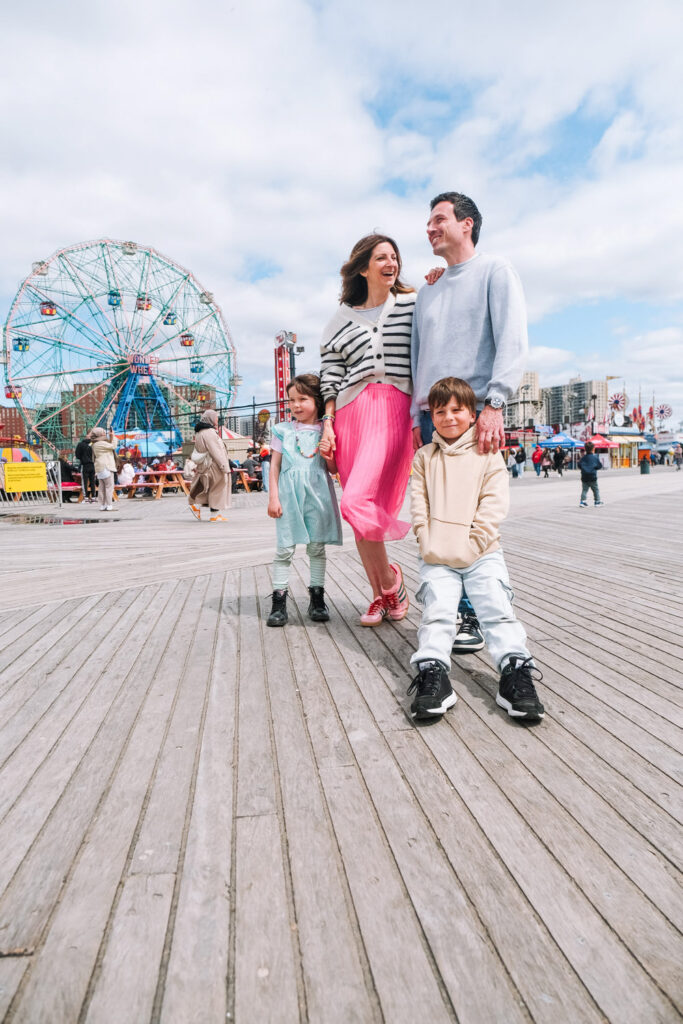 A family of four smiles and poses on a boardwalk, with a Ferris wheel and amusement park rides in the background under a partly cloudy sky—captured by a talented Brooklyn family photographer.