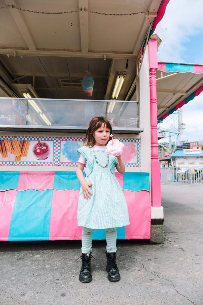 A young girl in a light blue dress and black boots stands in front of a colorful carnival stall, enjoying pink cotton candy on a sunny day—captured by a talented Brooklyn kids photographer.
