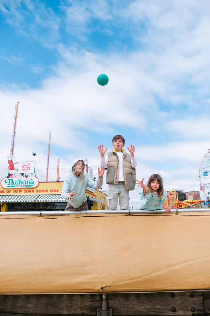 Three children stand behind a tan barrier at an amusement park, with one tossing a green ball into the air. Captured by a Brooklyn kids photographer, colorful carnival rides and signs, including Nathan’s, brighten the blue sky with clouds.