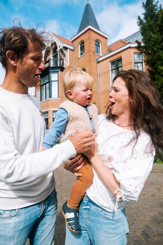 A man and woman stand outdoors in front of a brick building, holding a crying toddler between them. The concerned parents try to comfort their upset child during a candid family photography NYC session.