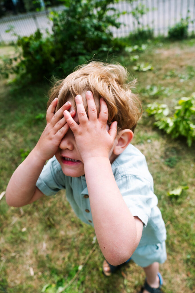 A young boy with light brown hair stands outside on grass, wearing a light blue shirt and shorts, covering his face with both hands as if playing or feeling shy—captured by a Brooklyn kids photographer against green bushes and a fence.