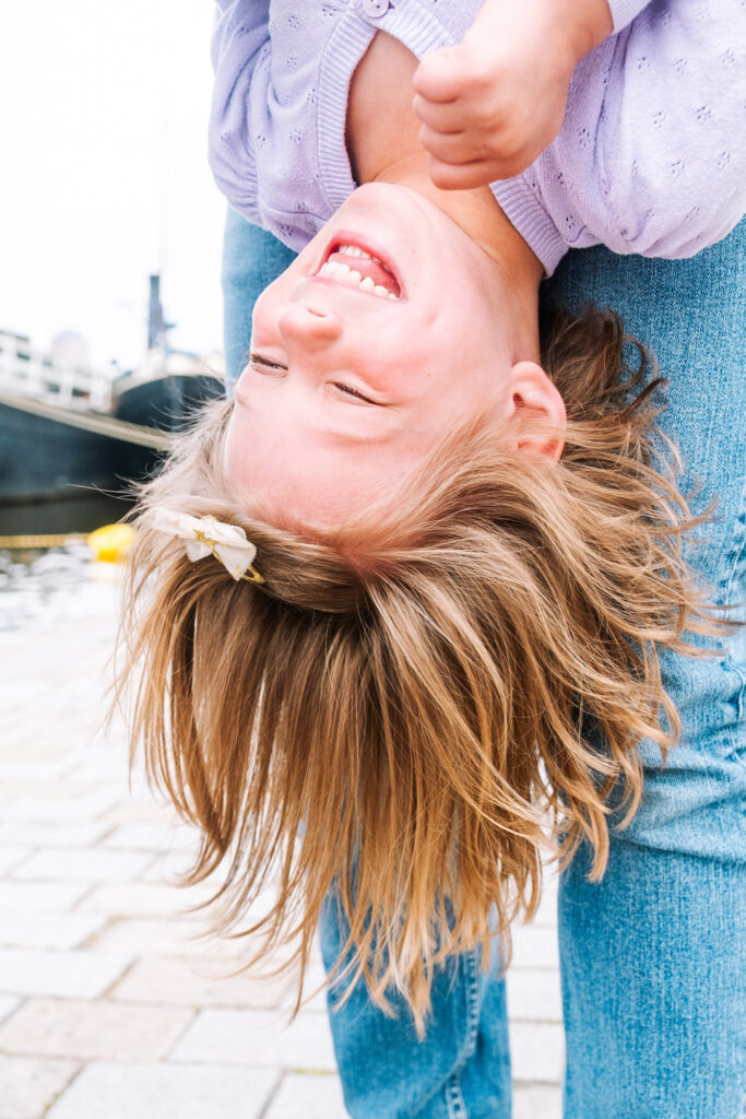 A young child with light brown hair and a white hair clip laughs joyfully while being held upside down outdoors near water and boats, captured in a light purple top and blue jeans by a talented Brooklyn kids photographer.